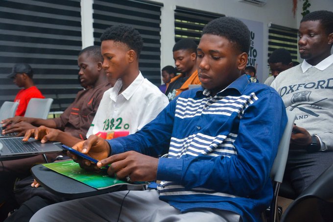 Photo of young men in a training or classroom setting, with one in the foreground wearing a blue striped shirt looking at a smartphone while others work on laptops.