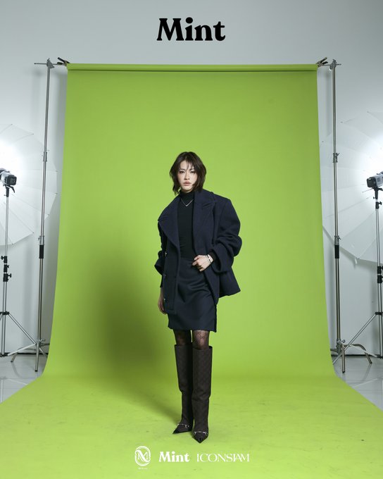 A woman poses in front of a bright green screen backdrop in a professional photo studio, wearing a dark oversized blazer, black skirt, patterned tights, and brown boots, with 'Mint' branding at the top and 'Mint ICONSiam' at the bottom.