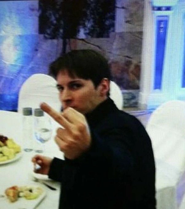 The image shows an adult male sitting at a dinner table with two wine glasses nearby, making peace signs with his fingers. He appears to be engaged in conversation or enjoying his meal as he makes these gestures. The surrounding setting includes chairs, dining tables, and other objects such as cups and bowls.