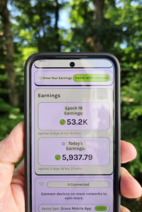 The image shows a person holding up their phone, which displays information about their earnings from a mobile app called "Grass". The screen of the phone is purple with green text that provides details about the user's progress in the app. The background features trees, suggesting an outdoor setting or park where the person has been working on their phone.