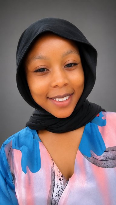 A smiling young woman wearing a black hijab and a colorful blue, pink, and purple patterned top poses against a gray background.