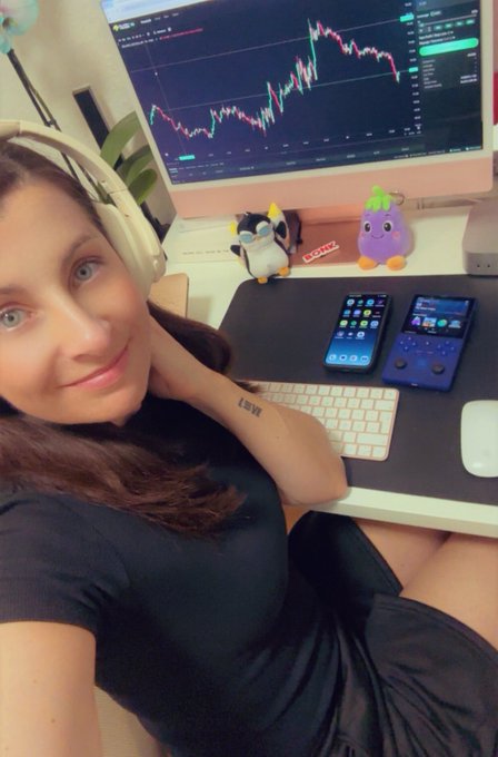 A woman with headphones takes a selfie at her desk, which features a computer monitor displaying a stock trading chart, a keyboard, two smartphones, and small plush toys.
