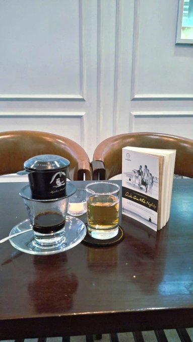A Vietnamese coffee setup on a dark wooden table, featuring a phin filter dripping coffee into a glass, accompanied by another glass of tea and a small book.
