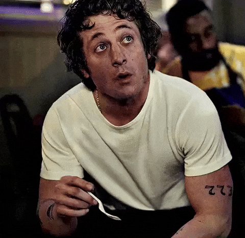 The image shows a man sitting at a table with his arm up, holding a spoon and eating food. There are other people present in the scene, but they do not seem to be paying attention to him or the spoon he's using. The focus of the image is on the man eating and the spoon, while the background appears to be unrelated to the main subject.