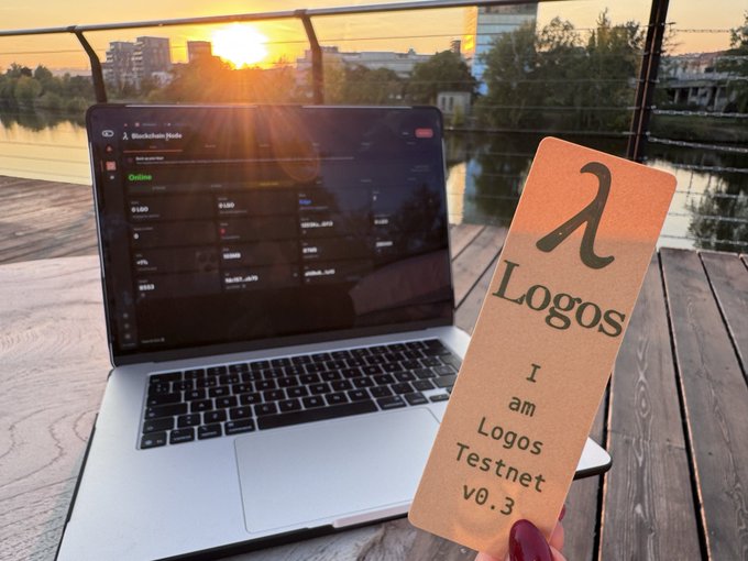 A laptop displaying a blockchain dashboard sits on a wooden table at sunset, while a hand holds a cardboard bookmark reading 'λ Logos - I am Logos Testnet v0.3'.