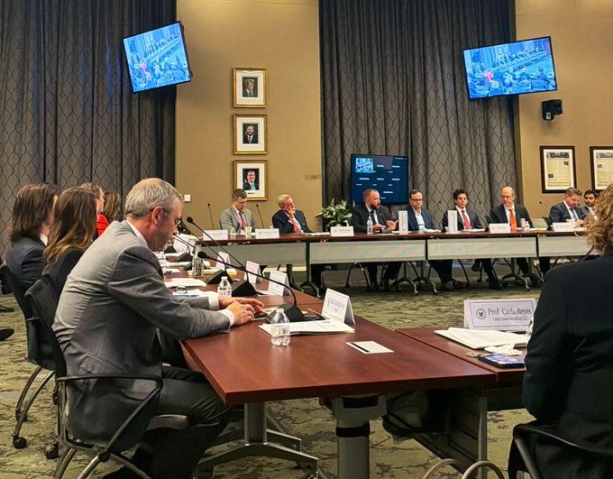 A group of people in business attire sit around a long conference table in a room with brown curtains and wall-mounted monitors, with a nameplate for 'Prof. Cipila Bayr' visible.