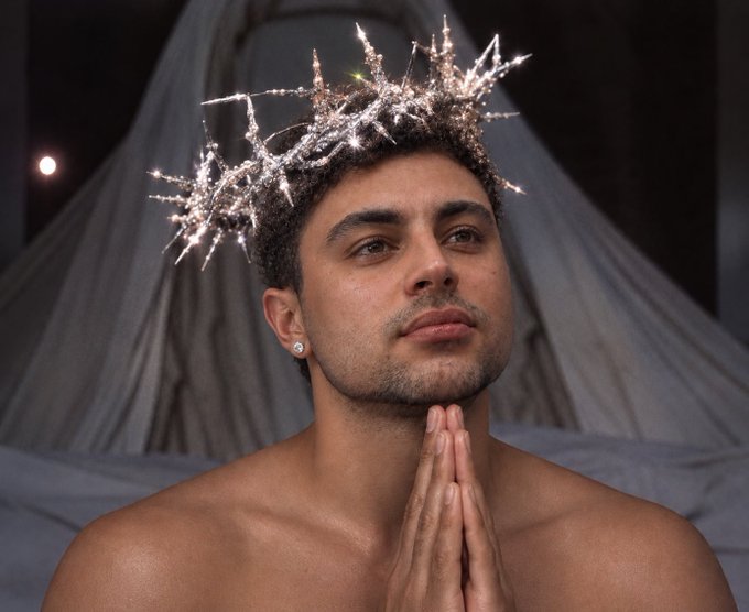A shirtless man with short dark hair wears a sparkling silver crown of thorns-like headpiece, holding his hands together in a prayer pose against a draped gray fabric background.