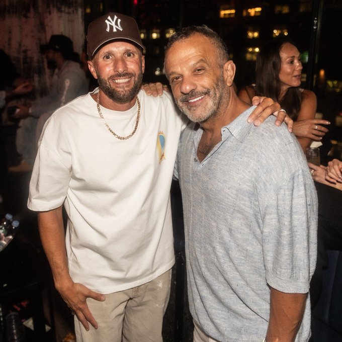 Two smiling men pose together at an event, one wearing a white t-shirt, gold chain, and NY Yankees cap, with the other in a light blue polo shirt with his arm around his companion.