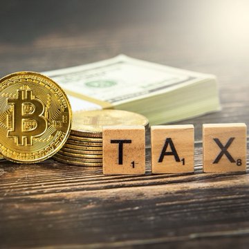 A gold Bitcoin coin and stack of coins sit next to wooden letter tiles spelling "TAX," with a banded stack of US dollar bills in the background. The image visually represents the new South African crypto tax framework, where digital assets like Bitcoin are treated as intangible assets subject to taxation rather than traditional currency.