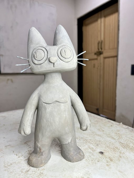 An unfinished gray clay sculpture of a cartoon-style cat character with large round eyes and whiskers, sitting on a white surface in a workshop.
