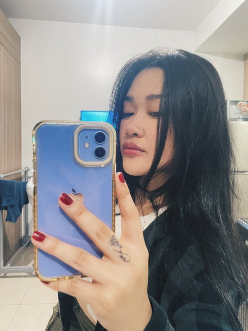 A mirror selfie of a woman with long dark hair and red nail polish holding a phone with a blue case, revealing a snake tattoo on her finger.