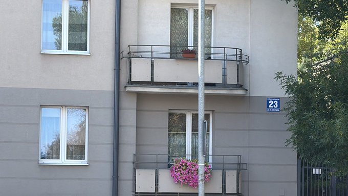 The exterior of a gray building with two balconies, a window with flowers on the lower balcony, and a blue sign reading "23".