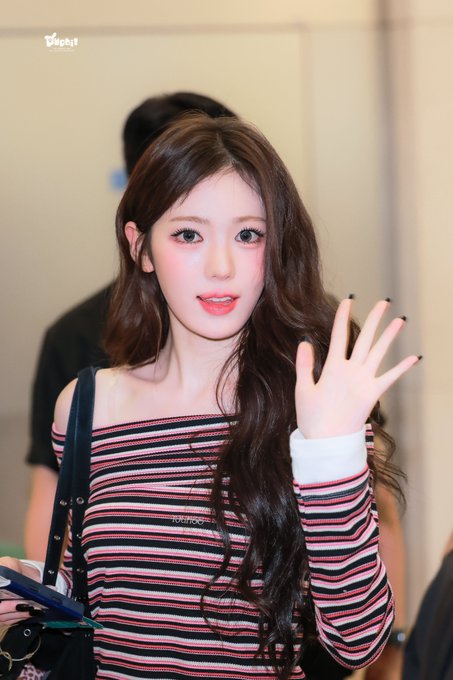 A young woman with long wavy brown hair and a pink-nude lip waves at the camera while wearing a black, red, and white striped off-shoulder top, captured in what appears to be an airport setting. The watermark "taepit" appears in the upper left corner, corresponding to the post's caption referencing SUI from KiiiKiii at ICN on August 22, 2026.