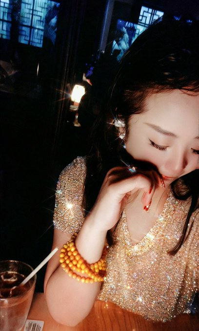 A woman in a sparkly gold dress poses at a dimly lit venue, touching her face while wearing a yellow beaded bracelet, with a drink visible in the foreground.