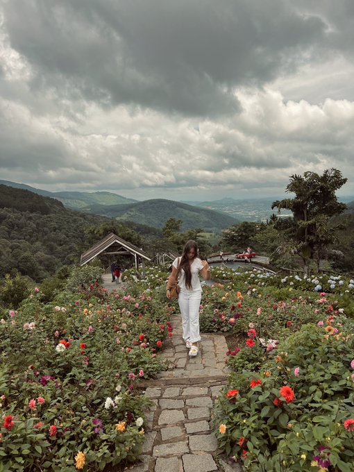 A person in white pants and a white shirt walks along a stone path through a colorful rose garden, with rolling green hills and dramatic cloudy skies in the background.