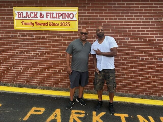 Two men pose in front of a brick wall beneath a "Black & Filipino - Family Owned Since 2025" sign, with "PARK" visible painted on the ground below, capturing their meeting at the family-owned beauty supply store in Detroit to discuss Pi Network and its strong Philippine community ties.