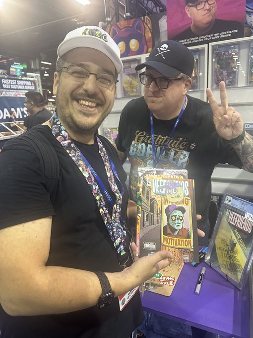 Two smiling men at what appears to be a convention; one holds a comic book titled 'NEVER TRIED IT AT COMIC-CON' with 'MOTIVATION' on the cover, while the other flashes a peace sign next to another comic book.