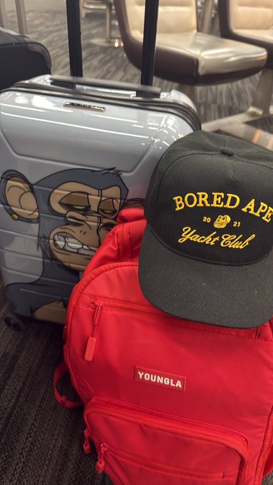 Travel gear including a silver suitcase featuring a Bored Ape character print, a red Youngla backpack, and a black 'Bored Ape Yacht Club 2021' cap sitting in an airport lounge.