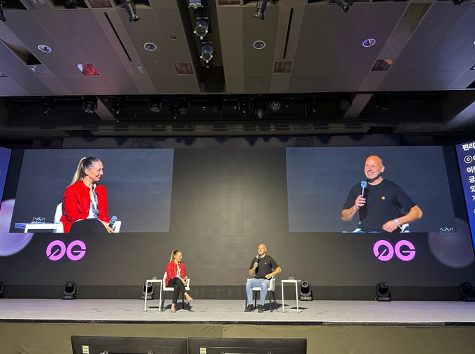 A man and woman sit on stage during a panel discussion at Korea Blockchain Week, with large screens behind them displaying their close-ups and the "DG" logo visible on stage. The image visually corresponds to the post by capturing the live moment featuring Ethereum co-founder Joe Lubin as he discusses the future of crypto adoption alongside institutional players.