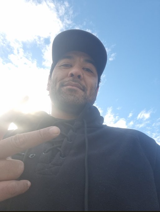 A selfie of a man wearing a black cap and dark hoodie, pointing his finger toward the camera against a bright, cloudy sky.