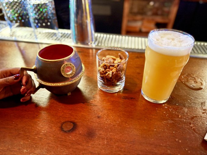 A person's hand with painted nails holds a decorative ceramic mug at a wooden bar, alongside a shot glass filled with candied nuts and a tall glass of beer with a frothy head. The polished bar surface reflects the warm lighting, fitting the post's playful mention of a "moll" enjoying date night drinks at a stylish spot with a bat hidden behind the bar.