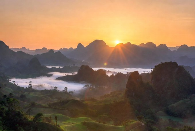 Sunrise over mist-covered karst mountains with rolling green hills in the foreground, matching the post's imagery of a sun awakening beyond distant peaks and lingering fog in the forest. The golden rays breaking through the silhouetted mountain range visually echo the poem's message of a peaceful, hopeful new day.