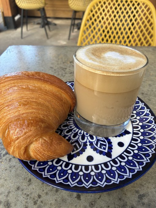 A golden croissant and a frothy latte are served on an ornate blue and white patterned plate atop a stone table, with yellow chairs visible in the background.