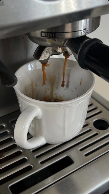 Espresso dripping from a stainless steel espresso machine portafilter into a small white ceramic cup sitting on a metal drip tray.