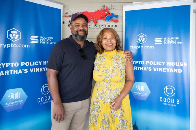 A man and a woman posing together in front of step-and-repeat banners for 'Crypto Policy House at Martha's Vineyard,' featuring logos for crypto.com, Solana Policy Institute, Blockchain Foundation, and Cocob Productions.