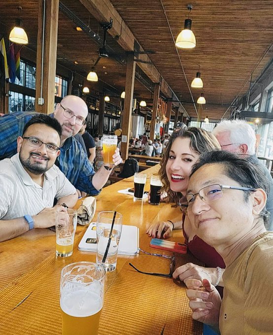 A group selfie of five people sitting at a long wooden table in a rustic restaurant or brewery with exposed wooden beams and hanging lights, smiling at the camera with beer glasses and drinks on the table.