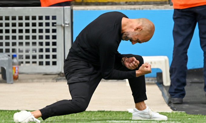 A bald man dressed in all black kneels on a soccer field sideline, pumping his fists in celebration while wearing white sneakers.
