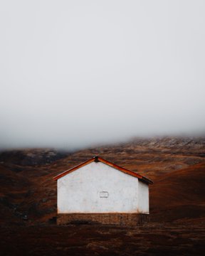 A small, white house with a red roof sits alone in a vast, barren, brown landscape under a foggy, pale sky.