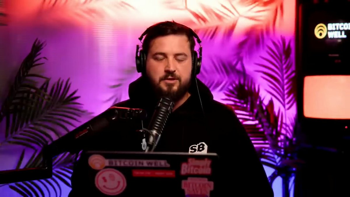 The image features a man wearing headphones while sitting at a desk with a laptop computer. The man appears to be singing into a microphone, possibly participating in an online radio show or podcast. The background of the image includes a purple wall and a TV screen displaying "bitcoin well".