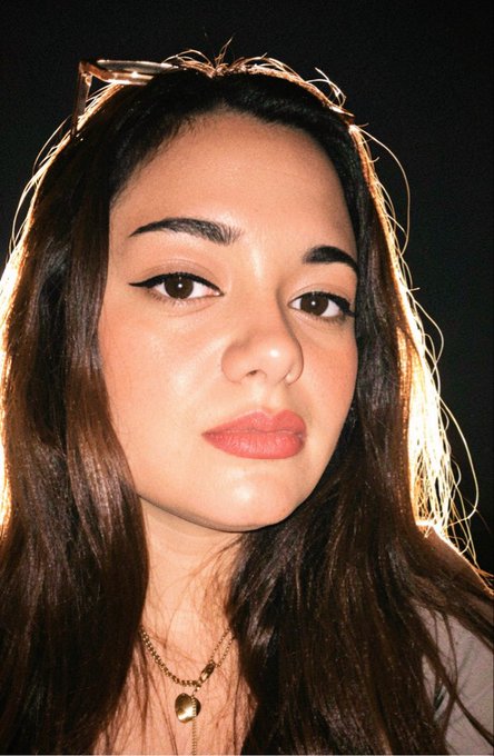A young woman with long dark hair, sunglasses perched on her head, and a heart pendant necklace poses for a close-up portrait against a dark background, styled in line with the "GM" (good morning) crypto greeting accompanying the post about blockchain infrastructure evolution.