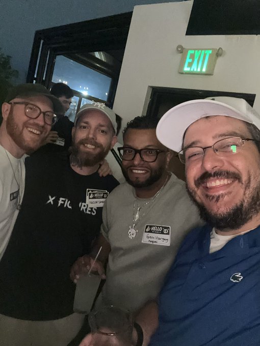 Four smiling men posing for a selfie at night near an illuminated EXIT sign; two are wearing name tags, with one reading 'Hello my name is Syrus Yturralde'.