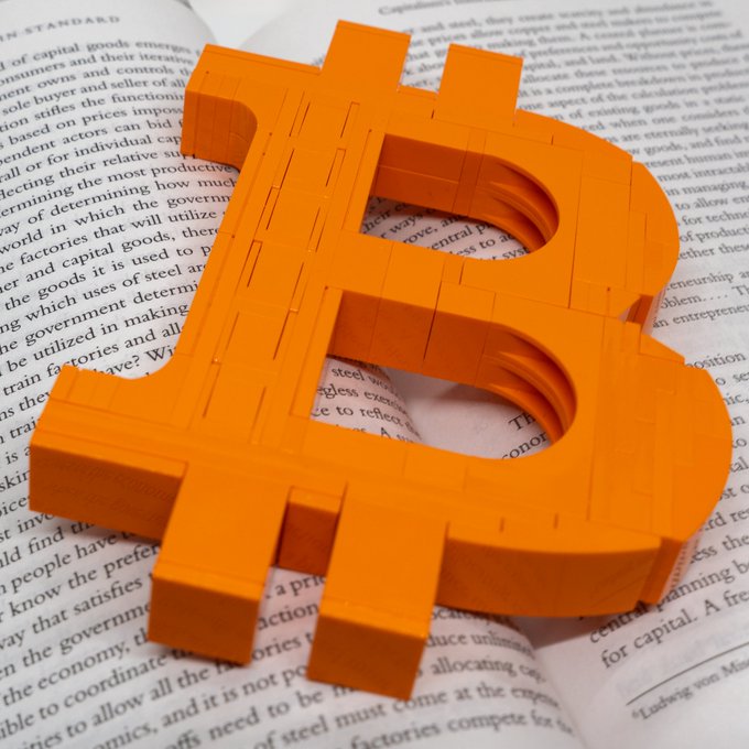 A 3D-rendered orange Bitcoin logo resting on the open pages of a book with visible text discussing economics and capitalism.