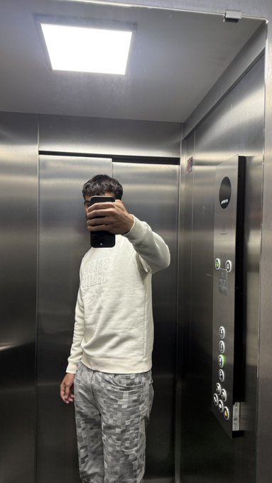 A person wearing a white sweatshirt and gray camouflage pants takes a mirror selfie with their phone inside a modern stainless steel elevator. The post reads "Good Morning Drop your $SOL address," suggesting the image is a casual, friendly morning greeting inviting others to share their Solana cryptocurrency wallet addresses.