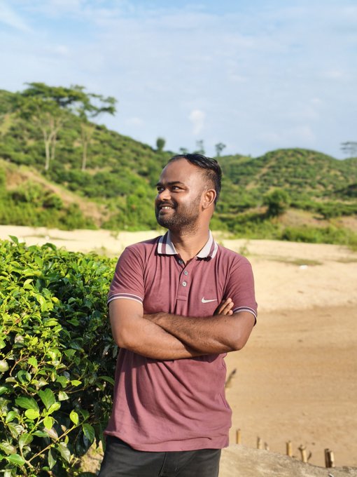 A man wearing a maroon polo shirt with his arms crossed, smiling while standing outdoors with green hills in the background.