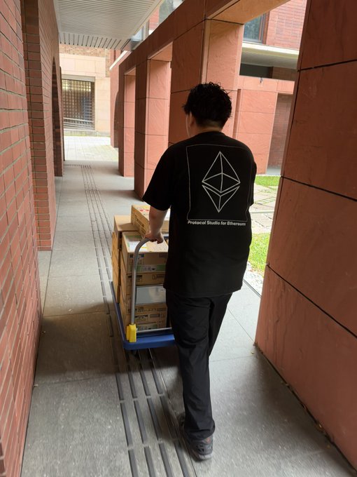 A person wearing a black 'Protocol Studio for Ethereum' t-shirt pushes a hand truck stacked with cardboard boxes down a brick corridor.