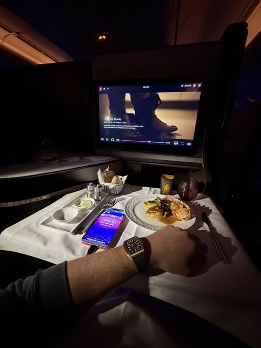 A passenger enjoys a first-class airline meal tray with a plated dish, appetizers, and a smartphone displaying crypto content, while watching a movie on the seatback screen, embodying the luxurious lifestyle associated with crypto wealth discussed in the post about buying $BNB after a market crash.
