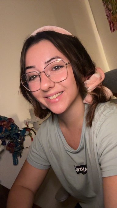 A selfie of a smiling young woman with short brown hair, glasses, wearing a pink headband with a bow and a gray t-shirt with reversed text reading "Live".