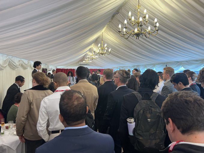 A crowded networking event inside a white marquee tent decorated with chandeliers, filled with attendees in business attire and backpacks mingling beneath the draped ceiling.