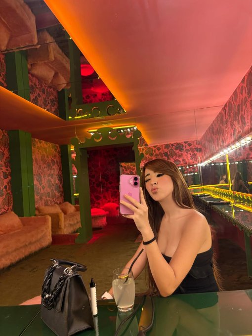 A young woman taking a mirror selfie with a pink iPhone in a vibrantly lit room with red and orange ceiling lights and patterned wallpaper; a drink and small bag sit on the green table beside her.