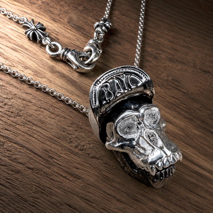 A close-up product photo of a silver skull pendant necklace with a crown inscribed 'BAD' resting on a wooden surface, featuring an ornate chain with clover-shaped links.