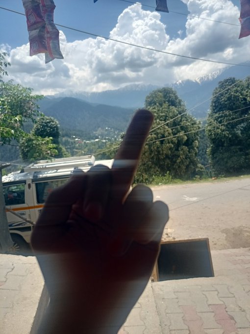 A photo taken through a window showing a hand with the middle finger raised in the foreground, with a mountainous landscape, trees, and a bus visible in the background.