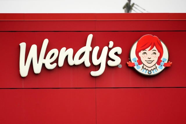 A Wendy's restaurant sign featuring the iconic red logo and the smiling red-haired mascot girl in a blue collar, mounted on a bright red wall. The image visually anchors the story of $WEN's surge, as the brand recognition and viral meme culture associated with Wendy's mascot helped fuel the stock's 25% spike and trading halt.