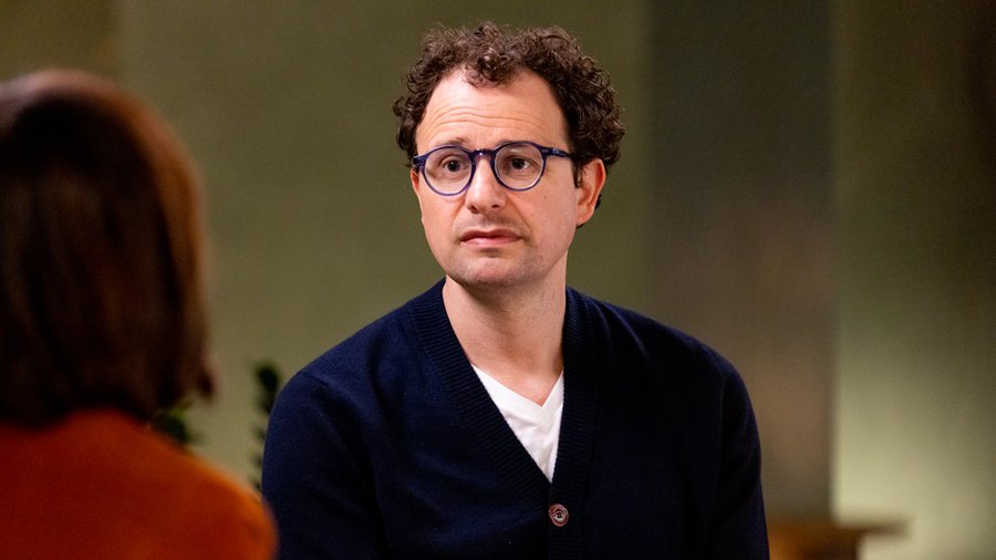 A curly-haired man wearing dark-rimmed glasses and a navy cardigan over a white shirt sits attentively across from another person with short brown hair, likely in an interview setting, with a contemplative expression on his face. The image appears to depict Dario Amodei, CEO of Anthropic, as referenced in the accompanying post about AI industry concerns and the potential risks of advanced artificial intelligence development.