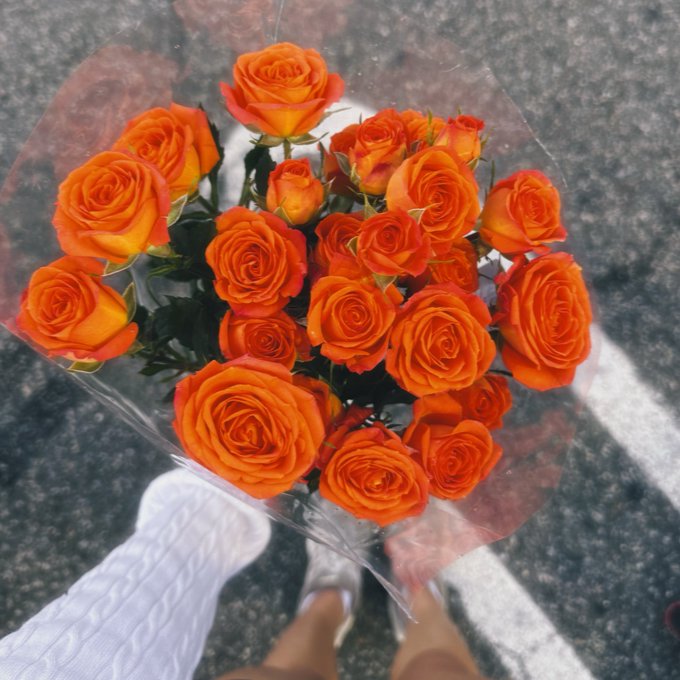 A bouquet of vibrant orange roses wrapped in clear cellophane, held above asphalt by a person wearing white sneakers.