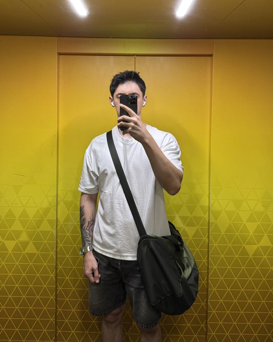 A young man takes a mirror selfie in an elevator with yellow walls, wearing a white t-shirt, dark denim shorts, and a crossbody bag, holding a phone in front of his face.