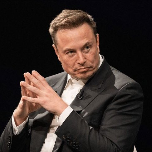 Elon Musk in a dark suit and white shirt, clasping his hands together with a serious expression.
